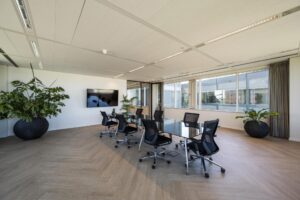 Moderne vergaderruimte aan de Thomas R. Malthusstraat met glazen tafel, ergonomische stoelen, grote planten en een flatscreen aan de muur.
